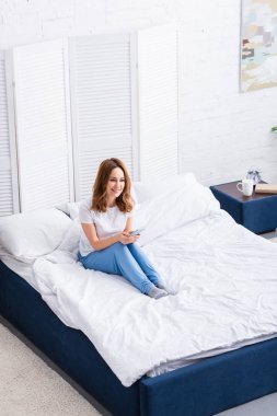 high angle view of attractive woman with ginger hair resting in bed and using smartphone at home on weekend clipart