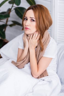 sick woman touching painful neck with scarf and sitting on bed at home and looking at camera clipart