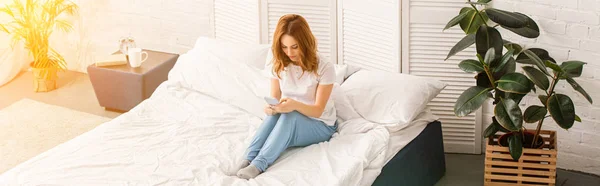 生姜髪のベッドで休んで 日光と自宅でスマート フォンを使用して魅力的な女性のパノラマ ビュー — ストック写真