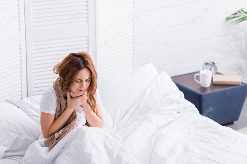 sick woman touching painful throat with scarf and sitting on bed at home