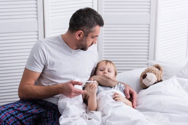 Baba yatak odasında hasta oğlu bakımı ve vücut ısısı termometresi ile denetleniyor