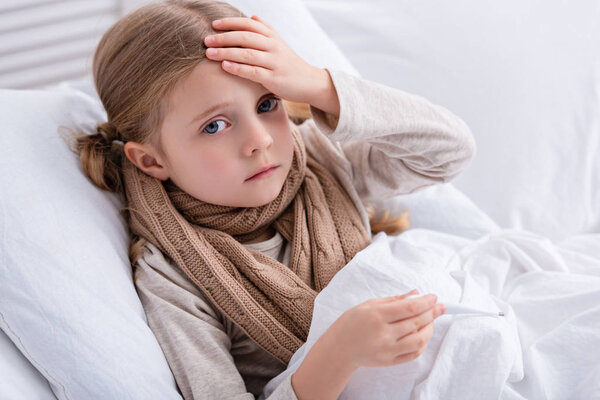 sick child with scarf over neck lying in bed, holding thermometer and touching forehead at home
