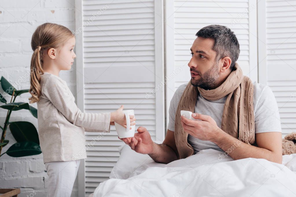 side view of adorable daughter giving cup of tea to sick dad in bedroom