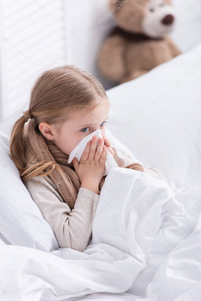 sick kid with scarf over neck lying in bed and blowing nose in tissue at home