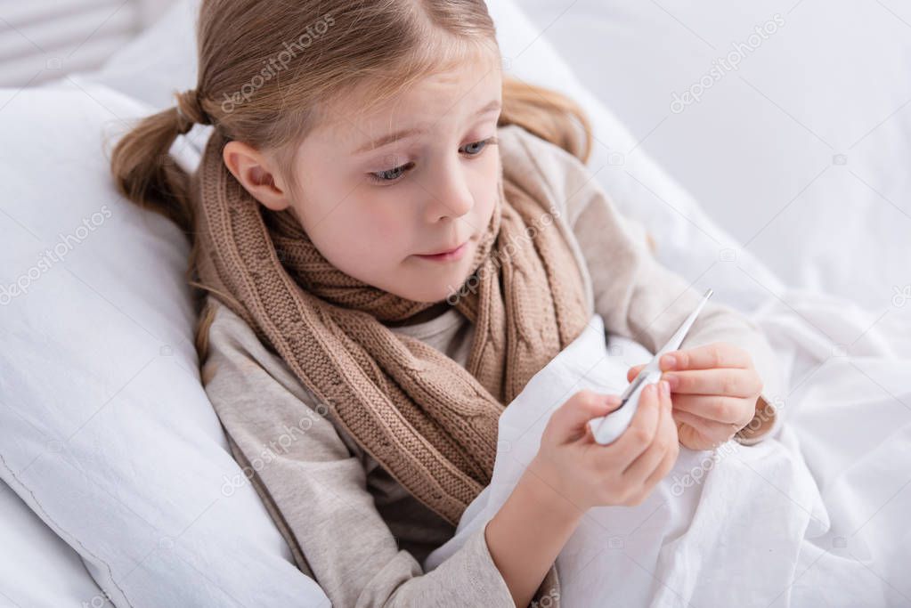 surprised sick child with scarf over neck lying in bed and checking temperature with thermometer at home