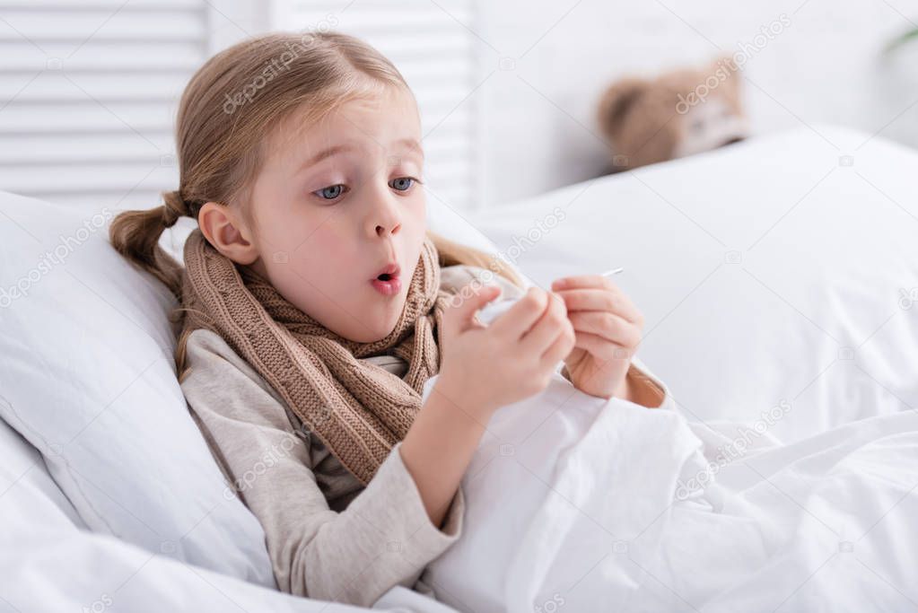 shocked sick child with scarf over neck lying in bed and checking temperature with thermometer at home