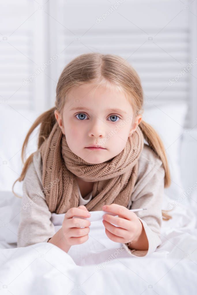 sick child with scarf over neck lying in bed, holding thermometer and looking at camera at home