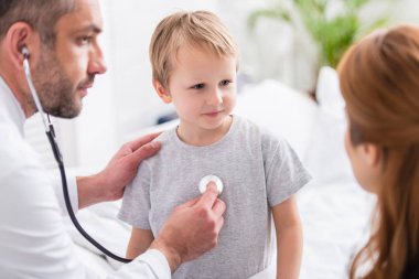 selective focus of pediatrist in white coat examining sick boy with stethoscope clipart