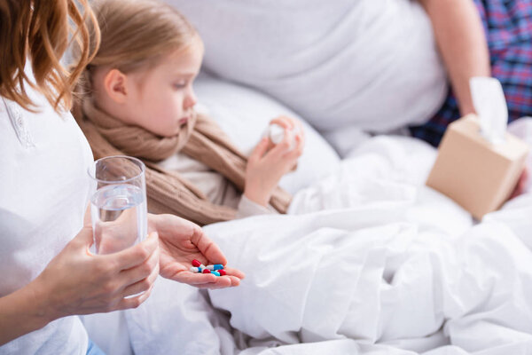 cropped image of parents taking care of sick daughter in bedroom, giving her pills and glass of water