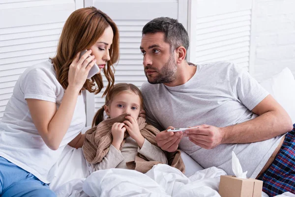 mom calling doctor and dad checking temperature of sick daughter in bedroom