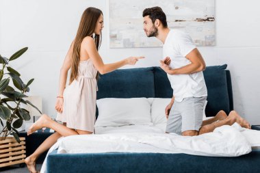 young attractive woman poiting finger angrily on stressed gesturing man while standing on bed clipart