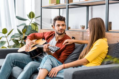 young handsome man sitting on sofa in living room and playing acoustic guitar for girlfriend clipart