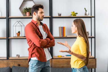 angry boyfriend standing with crossed arms while girlfriend quarreling in living room clipart