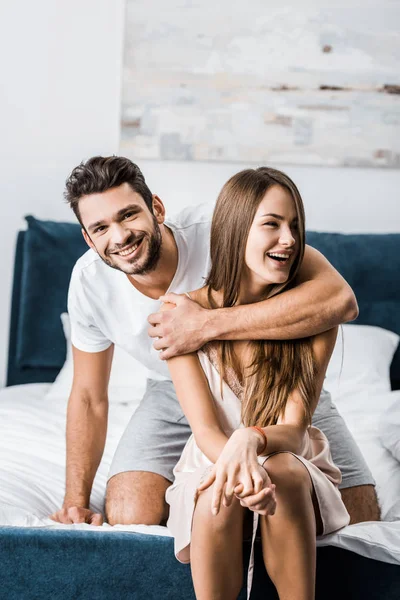 Jovem Abraçando Mulher Com Uma Mão Sorrindo Enquanto Sentado Cama — Fotografia de Stock Grátis