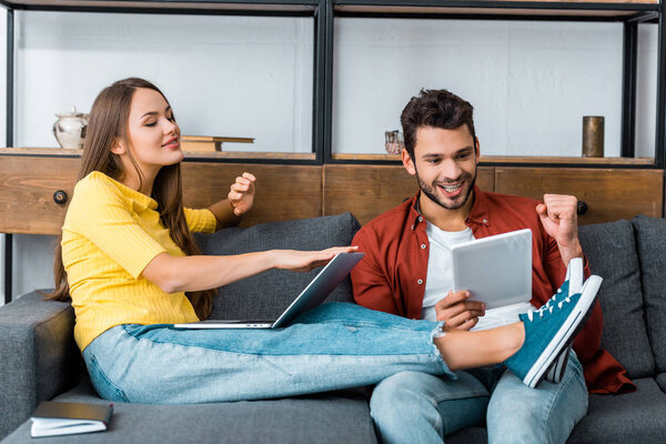 young cheerful couple spending time together on sofa with laptop and digital tablet