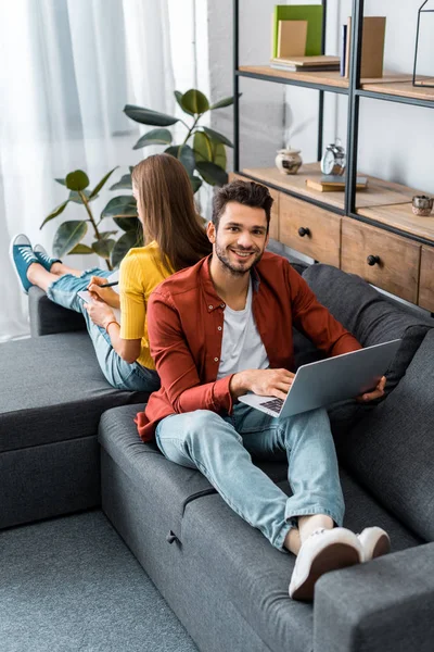 Joven Hombre Sonriente Sentado Sofá Celebración Portátil Espalda Con Espalda — Foto de stock gratis