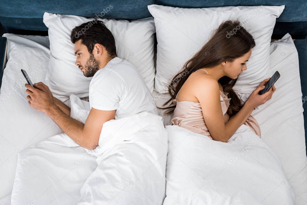 top view of young couple lying in bed in morning and using smartphones
