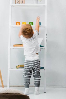 cute toddler boy standing near rack and reaching for toys on shelves clipart