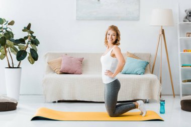 woman in sportswear kneeling on yellow fitness mat, looking at camera and smiling in living room