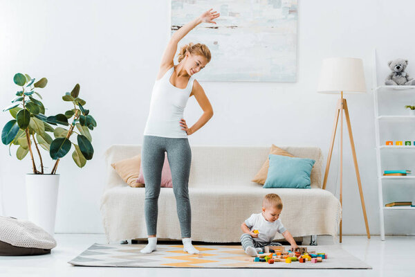 smiling woman doing exercise and looking at cute toddler boy playing with multicolored cubes on carpet in living room