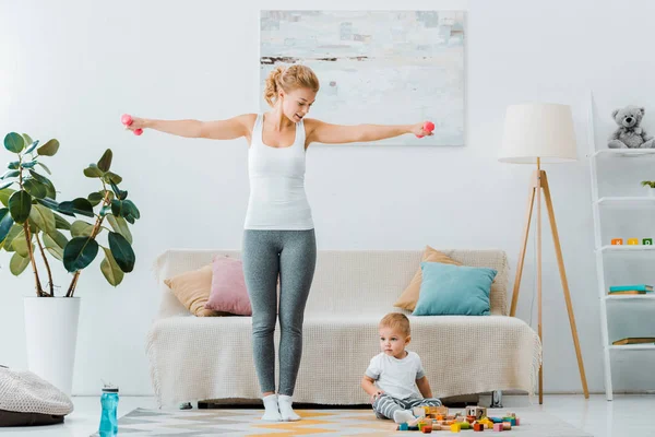 Beautiful Woman Holding Dumbbells Looking Toddler Boy Sitting Carpet Multicolored — Stock Photo, Image