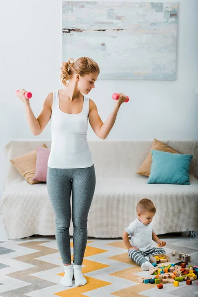 Atractiva Mujer Haciendo Ejercicio Con Pesas Mirando Niño Sentado Alfombra — Foto de Stock