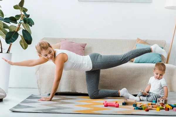 Mujer Haciendo Ejercicio Cerca Del Sofá Mirando Niño Jugando Con — Foto de Stock