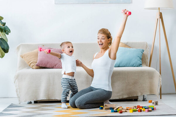 beautiful mother and cute toddler son holding dumbbells in raising hands and laughing in living room