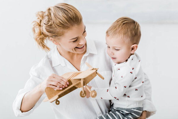 smiling woman holding wooden plane model and playing with cute toddler boy at home
