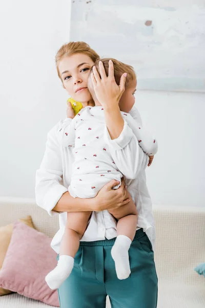 Mutter Hält Kleinkind Arm Und Blickt Wohnzimmer Kamera — kostenloses Stockfoto