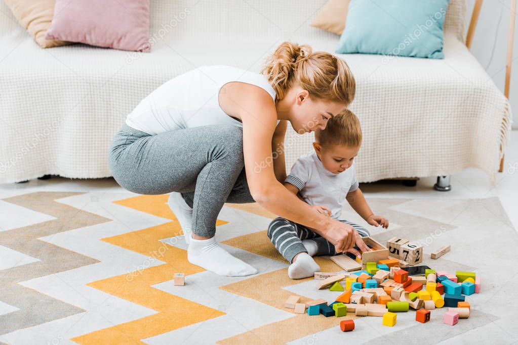 attractive woman and cute toddler boy playing with multicolored wooden cubes on carpet at home
