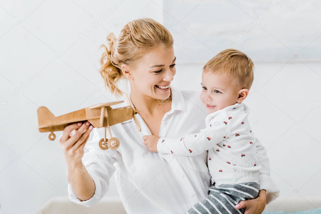 smiling woman holding wooden plane model and playing with adorable toddler boy at home