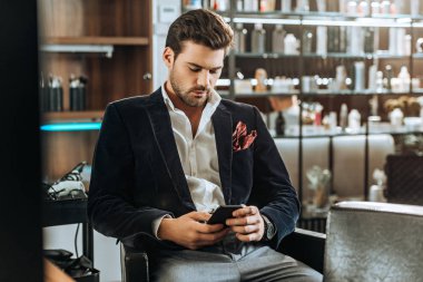 stylish handsome young man using smartphone while sitting in beauty salon clipart