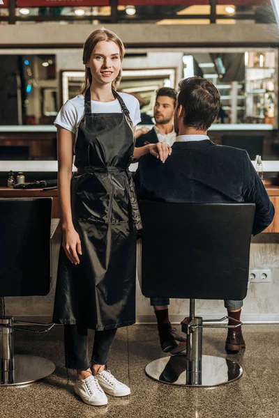 美容室で椅子に座っている男性のクライアントしながらカメラに笑顔美しい若い美容師 — ストック写真
