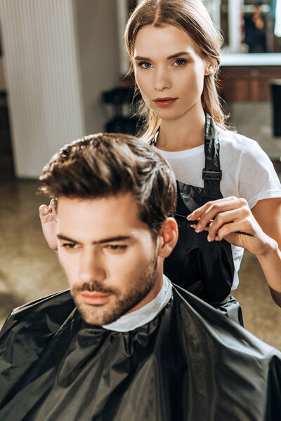 hairstylist looking at camera while cutting hair to handsome young man in beauty salon 