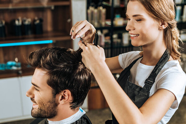 smiling young hairdresser cutting hair to handsome happy man in beauty salon