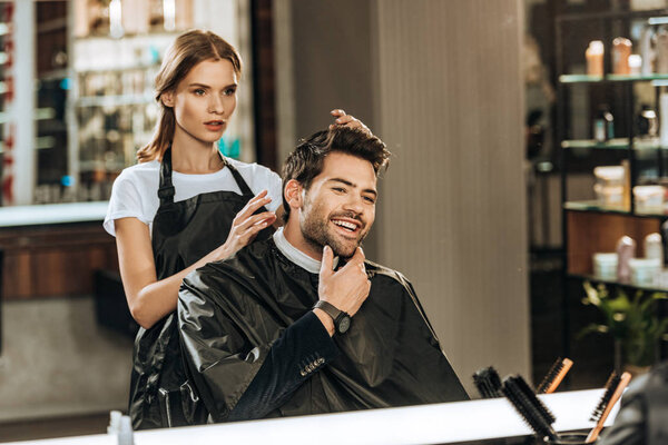 young female hairstylist and handsome smiling man looking at mirror in beauty salon