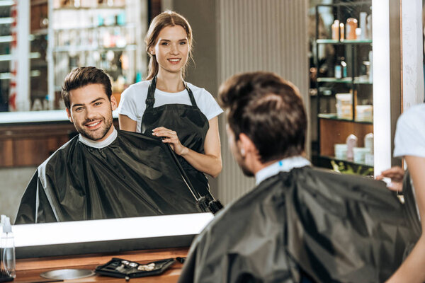 smiling young hairstylist and handsome client looking at mirror in beauty salon