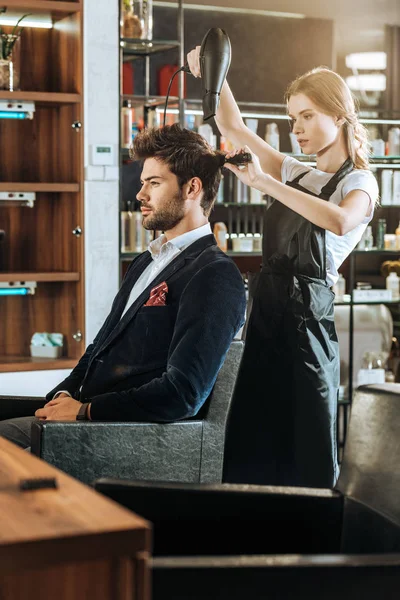 在美容院美丽的年轻发型师梳理和干燥头发英俊的客户的侧视图 — 图库照片