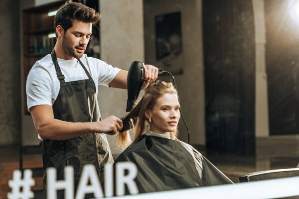 reflection in mirror of hairstylist drying hair to beautiful young woman in beauty salon 