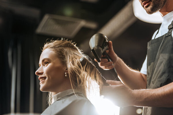 low angle view of hairstylist combing and drying hair to beautiful young woman in beauty salon