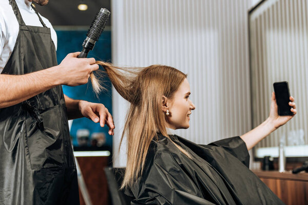 side view of smiling girl taking selfie with smartphone while hairstylist doing haircut in beauty salon