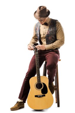 young mixed race male musician in hat and eyeglasses tuning acoustic guitar while sitting on chair isolated on white clipart