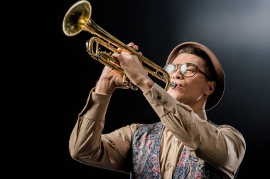 young male jazzman in hat and eyeglasses playing on trumpet isolated on black clipart