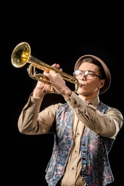 handsome male jazzman in hat and eyeglasses playing on trumpet isolated on black clipart