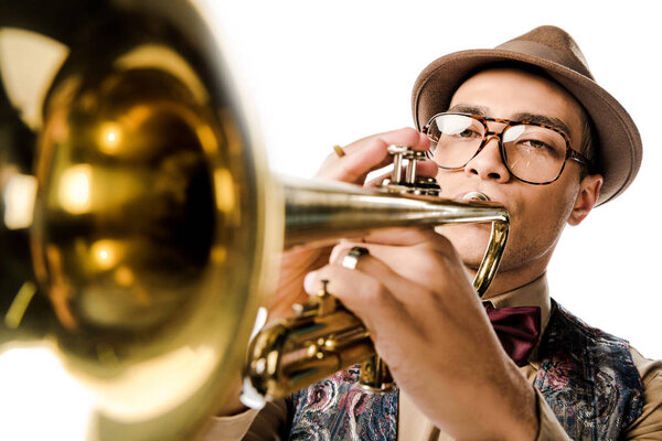 selective focus of young mixed race man in stylish hat and eyeglasses playing on trumpet isolated on white 