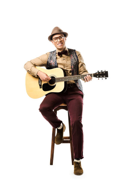cheerful mixed race male musician in hat and eyeglasses playing on acoustic guitar while sitting on chair isolated on white