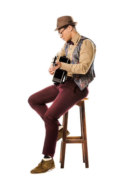 side view of mixed race male musician in hat and eyeglasses playing on acoustic guitar while sitting on chair isolated on white