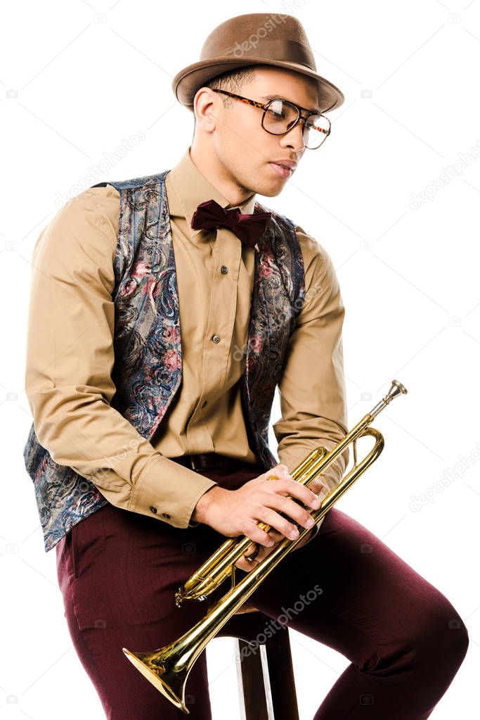 serious mixed race male jazzman holding trumpet and sitting on chair isolated on white