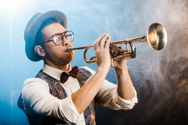 şapka ve gözlük trompet üzerinde dramatik aydınlatma ve duman ile sahnede oynarken şık jazzman